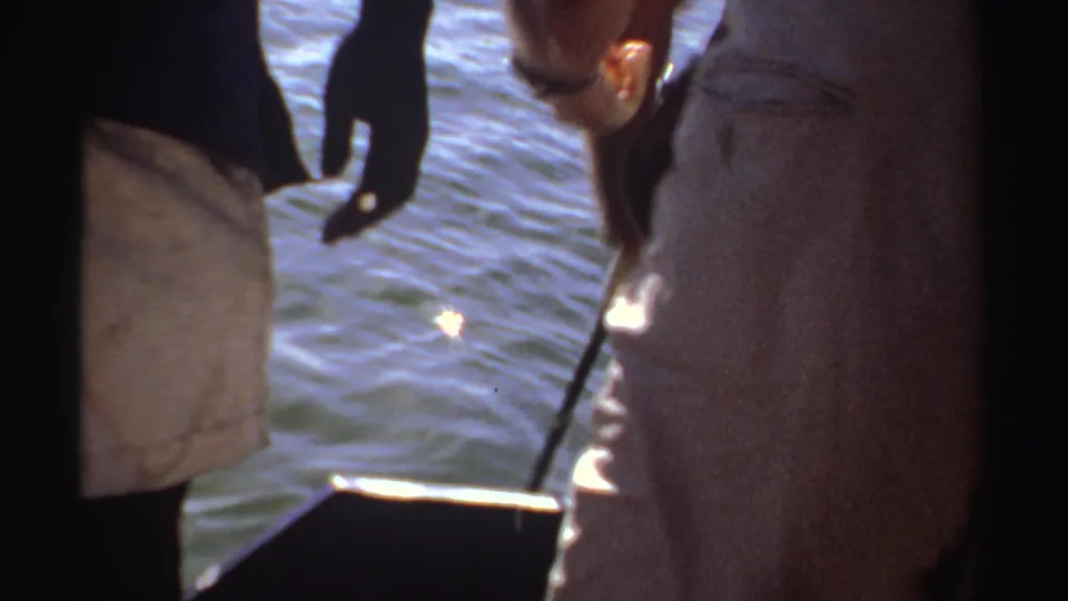 1969 Sudan frame of adult men on a small boat, with one bending near the side and bright water behind them.