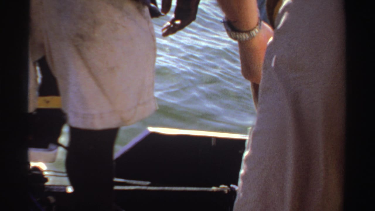 1969 Sudan 8mm frame with a cropped view of a person seated or standing near a boat edge, white cloth, hands, and water behind.
