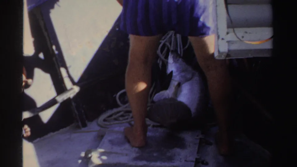 1969 Sudan 8mm frame showing a barefoot adult standing on a boat deck near rope and rail, with a silhouetted figure at right.