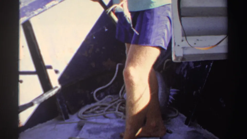 1969 Sudan home-movie frame with an adult moving across a boat deck beside coiled rope and a fish in sharp sunlight.
