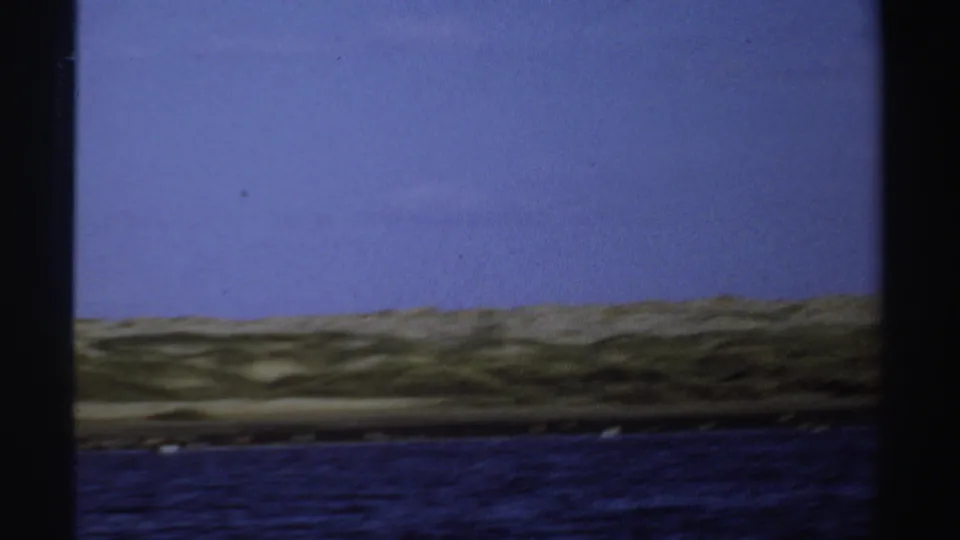 1969 Sudan 8mm frame showing rippled water, a low sandy shoreline, pale sky, and contact-sheet borders with birds no longer prominent.