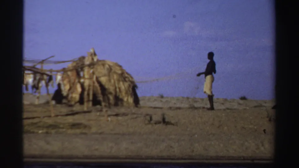 1969 Sudan riverside frame with a man walking past a thatched shelter and pole drying rack in open terrain.