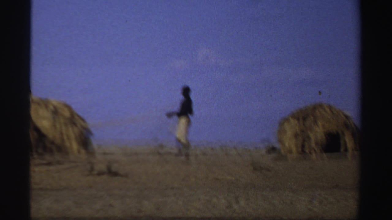 Distant person near thatched huts on a sandy riverbank under a blue sky.