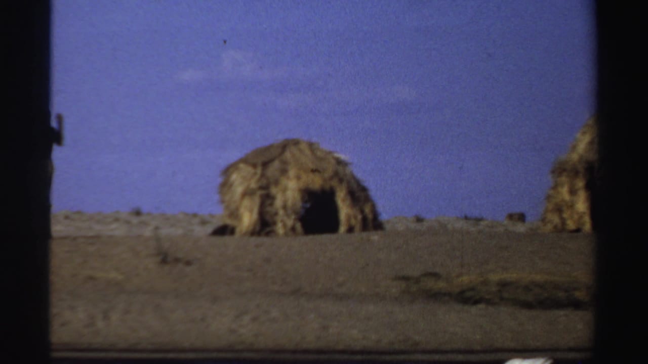 1969 Sudan frame showing thatched huts with dark entrances on a dry riverbank landscape.