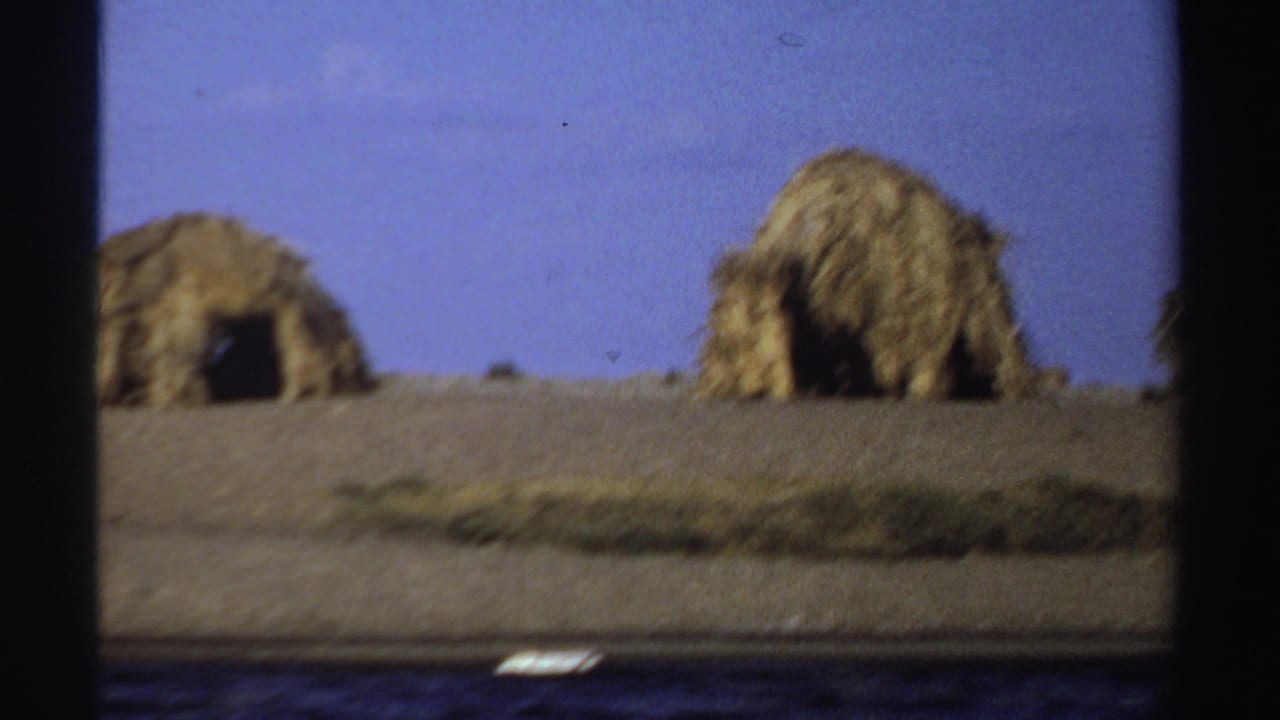 1969 Sudan home-movie frame with thatched huts along dry ground above a visible river edge.