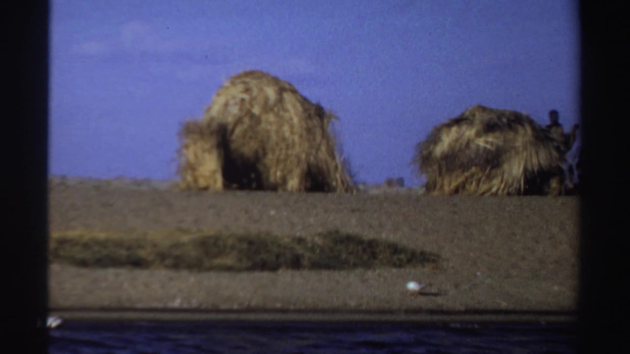 1969 Sudan frame showing thatched huts on dry riverbank terrain, with a person partly visible at the right edge.