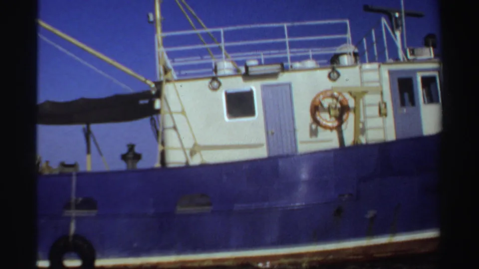 1969 Sudan 8mm frame of a blue boat with a white cabin, deck railings, ropes, and an orange life preserver.