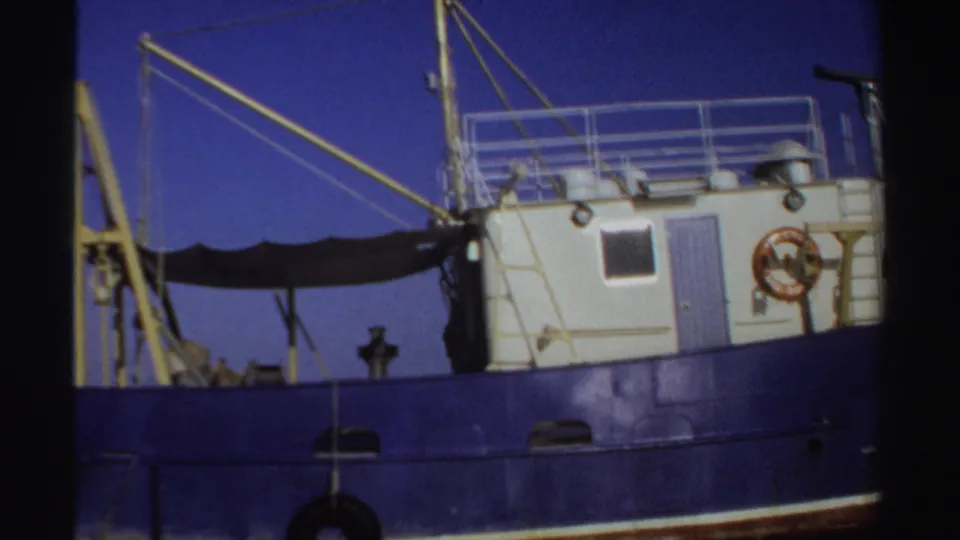 1969 Sudan 8mm frame showing a blue working boat with white cabin, yellow crane, ropes, and railings under clear sky.
