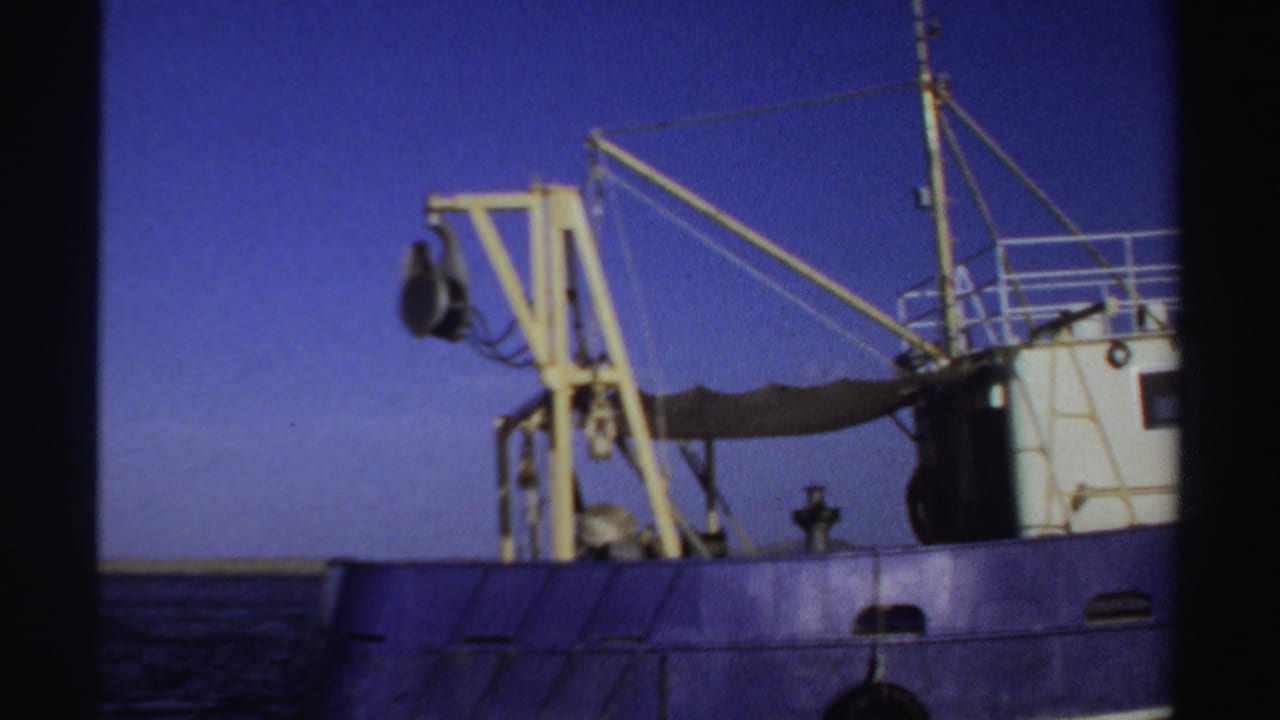 1969 Sudan 8mm frame showing the side of a blue industrial fishing boat with a yellow crane, rigging, and calm sea behind it.