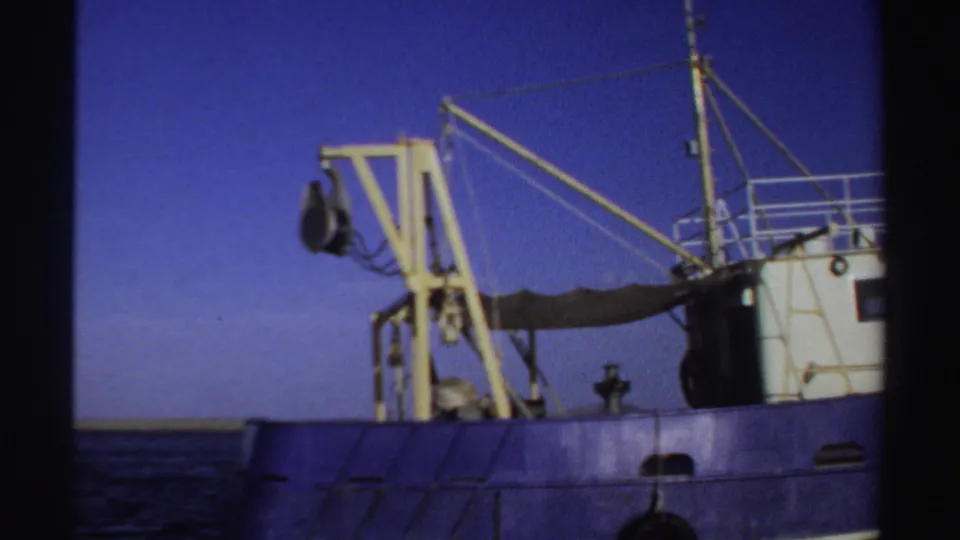 1969 Sudan 8mm frame showing the side of a blue industrial fishing boat with a yellow crane, rigging, and calm sea behind it.