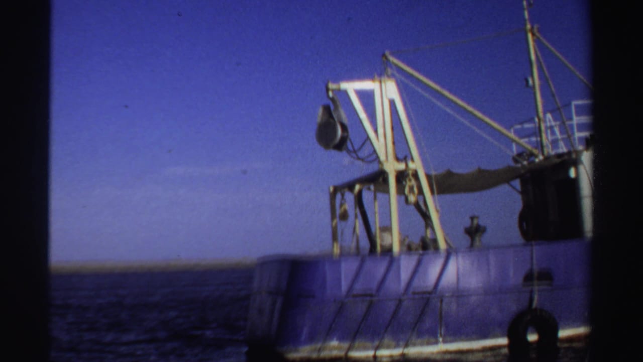 1969 Sudan 8mm frame with a blue industrial fishing vessel, yellow crane, rigging, tire fender, and distant low horizon.
