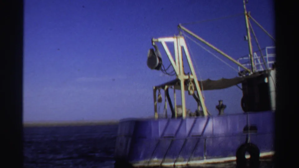 1969 Sudan 8mm frame with a blue industrial fishing vessel, yellow crane, rigging, tire fender, and distant low horizon.