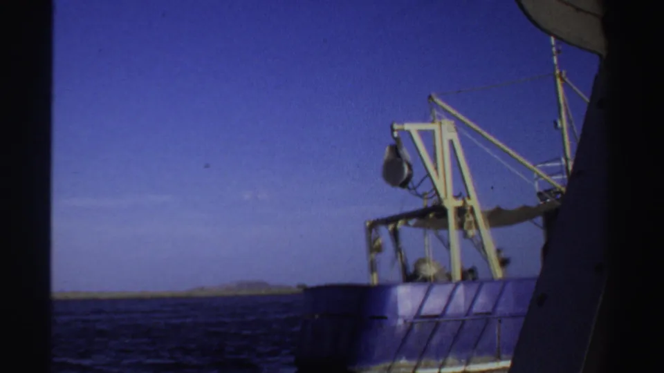1969 Sudan 8mm frame showing the blue fishing boat partly obscured by the filming vessel, with open water and distant low land.