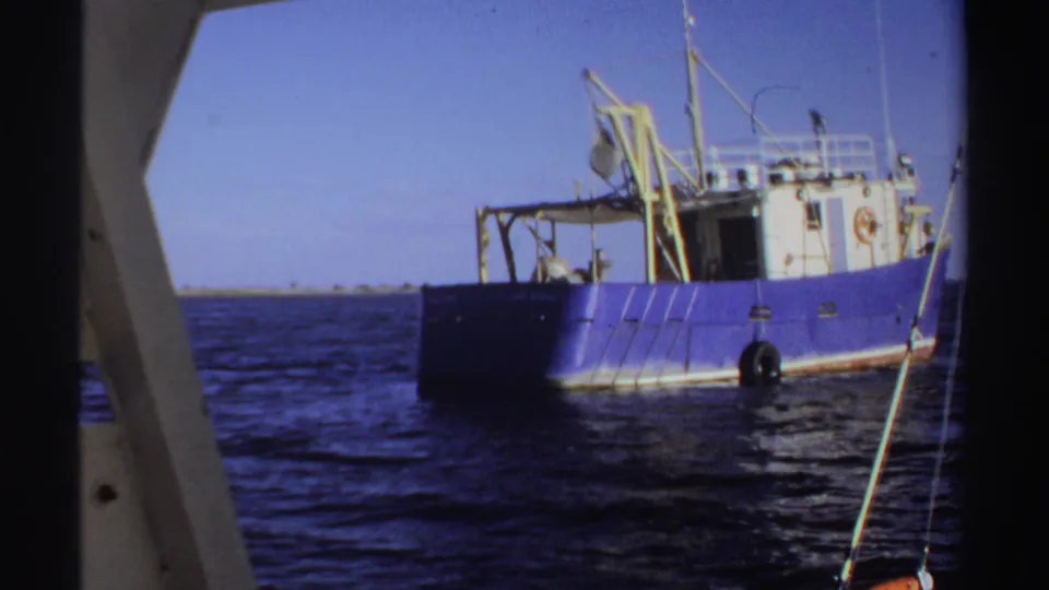 1969 Sudan 8mm frame showing the blue industrial fishing boat at a wider distance with wheelhouse, crane, rigging, and tire fenders visible.