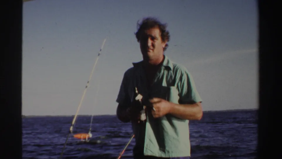 1969 Sudan 8mm frame showing an adult man on deck holding a bottle, with fishing rods and water behind him.