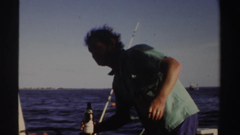 1969 Sudan 8mm frame with an adult man leaning on deck near a bottle, fishing rods, and open water.