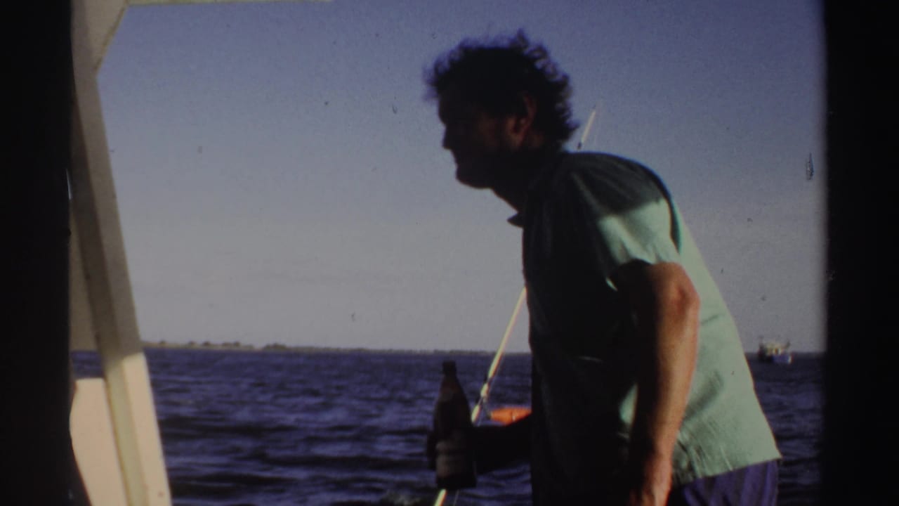 In Sudan-associated 1969 8mm footage, a man stands in profile on a boat holding a beer bottle beside open water.