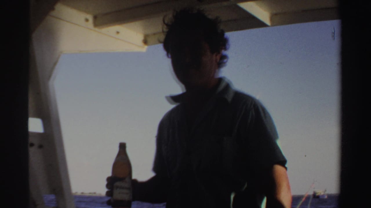A man on a boat in Sudan-associated 1969 home-movie footage holds a beer bottle while facing the camera in partial shadow.