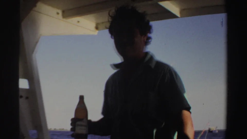 A man on a boat in Sudan-associated 1969 home-movie footage holds a beer bottle while facing the camera in partial shadow.