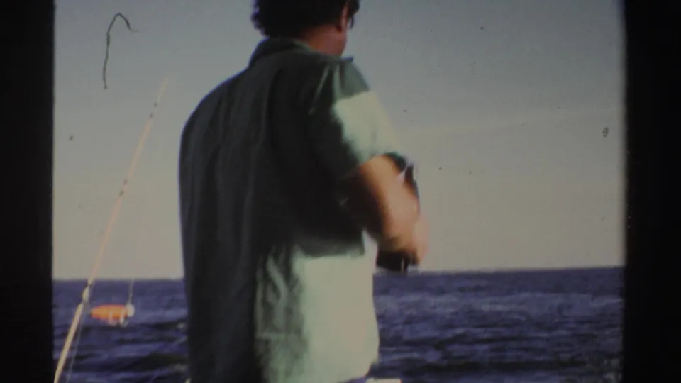 A man is seen from behind on a boat in Sudan-associated 1969 home-movie footage, with fishing rods and open water visible.