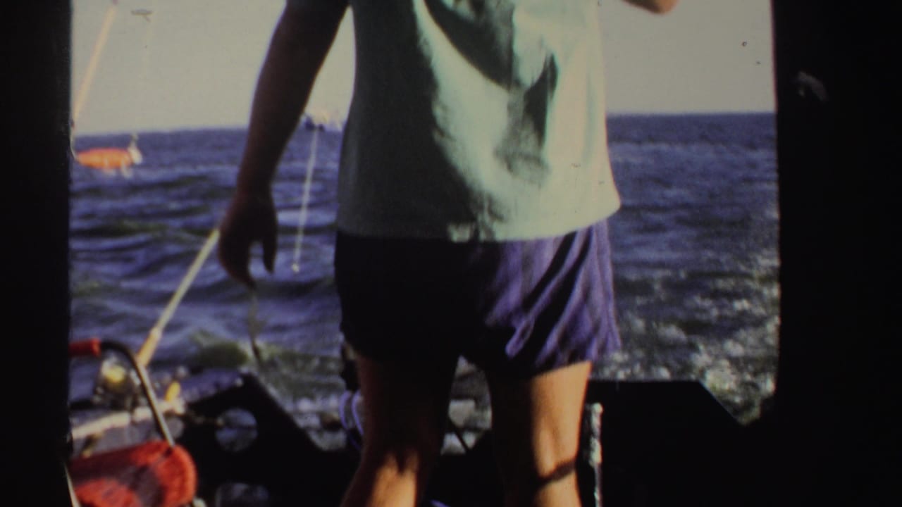 In Sudan 1969 footage, a man in a pale shirt and shorts stands on a boat with water and deck equipment behind him.
