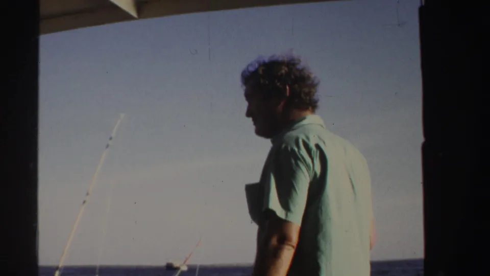 A man in a light green shirt stands in profile on a boat in Sudan, with fishing rods, horizon, and open water behind him.