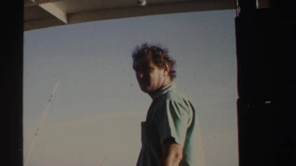The man turns toward the camera in the 1969 Sudan home-movie shot, with fishing rods and pale sky behind him.