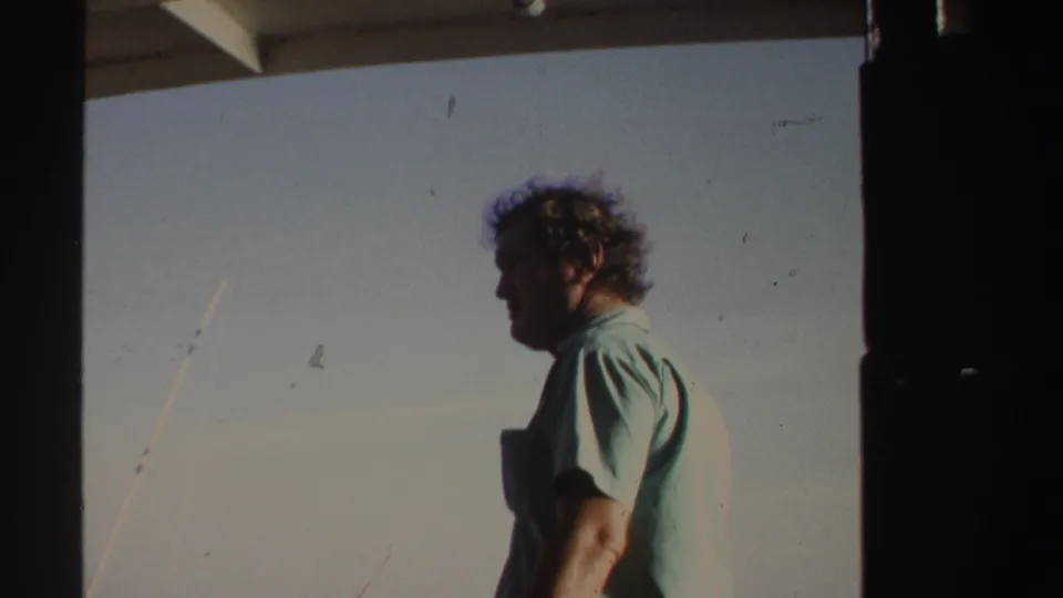 A man in a light green shirt looks away again on a boat in Sudan, framed by dark structure with water and sky behind him.