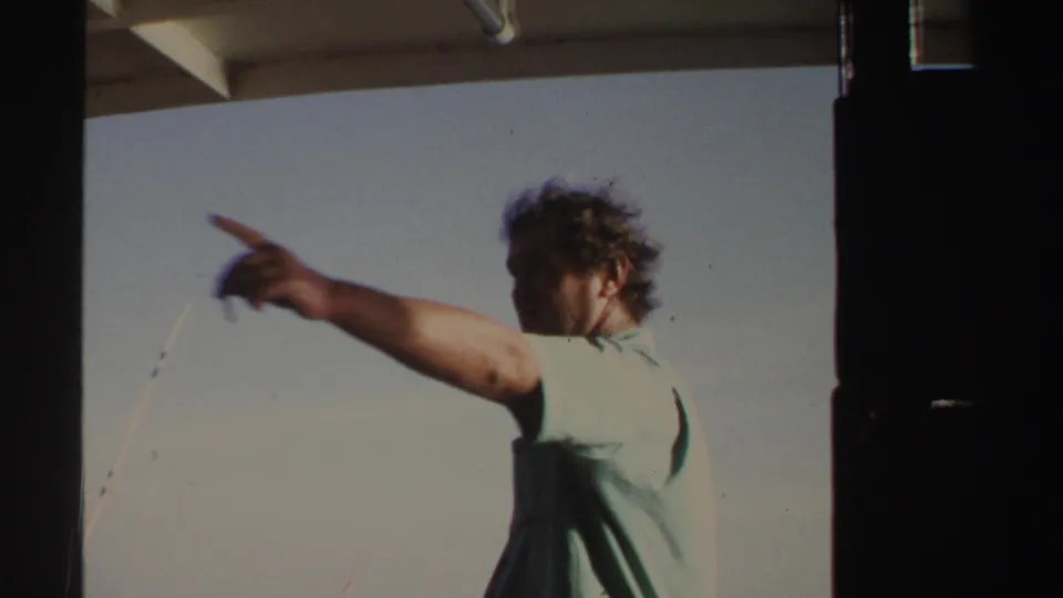 In Sudan 1969 home-movie footage, an adult man in a light blue shirt points outward from a boat against open sky.