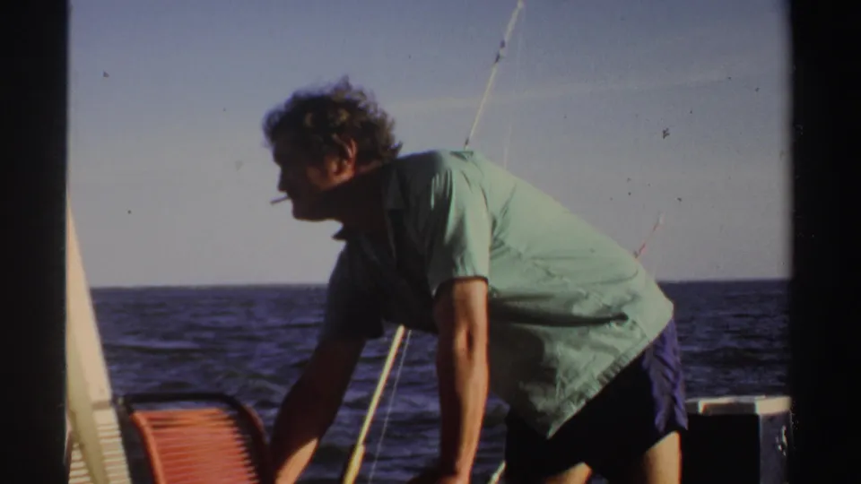 The man leans forward on the boat in Sudan in 1969, with fishing rods, railing, and open water visible behind him.