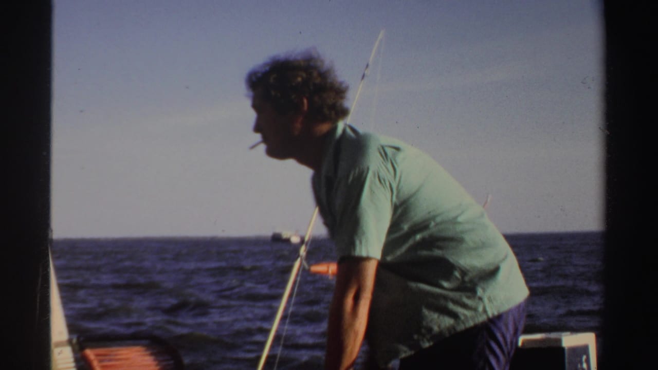 Man in a light green shirt leans on a boat beside fishing rods on open water.