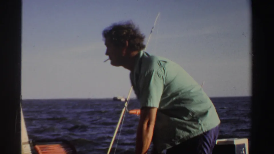 In 1969 Sudan home-movie footage, an adult man with a cigarette leans forward on a boat with fishing rods against open water.