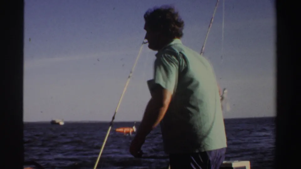 A side view shows the adult man standing near a fishing rod on a boat, with dark water and a distant horizon visible.