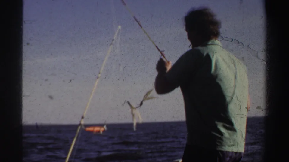 Seen from behind, the man handles a fishing rod on the boat while the waterline and clear sky remain in view.