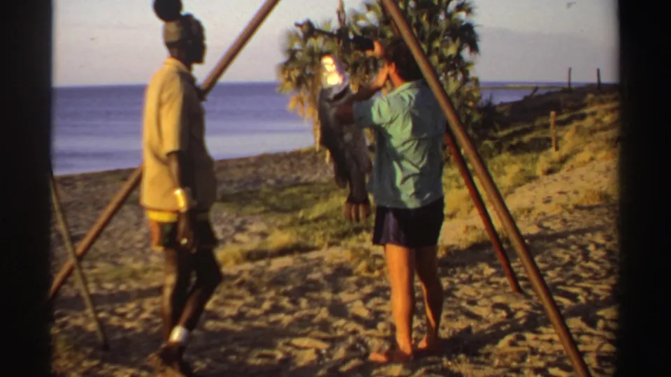 1969 Sudan 8mm frame showing two men and a large fish hanging from a pole against a beach and sea backdrop.