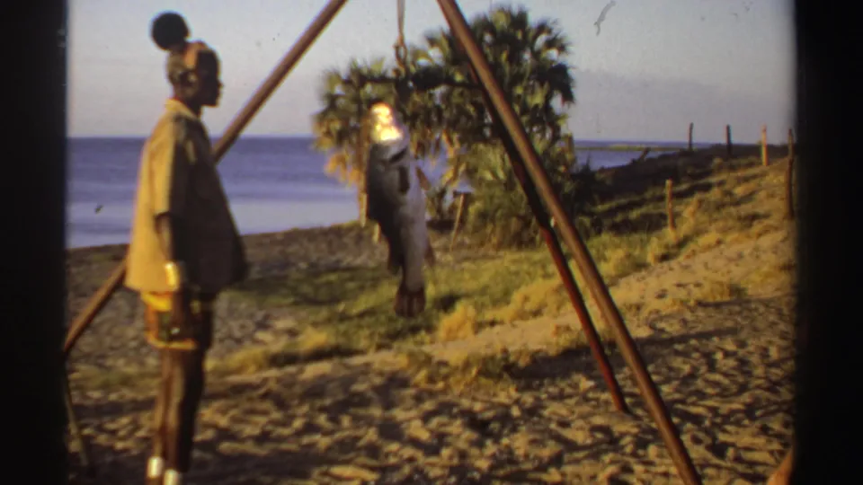 1969 Sudan coastal frame with a man standing by a hanging fish, palm vegetation, wooden poles, sand, and sea visible behind.