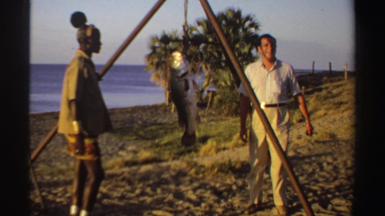 Two men stand on a beach beside a large fish hanging from a wooden tripod.