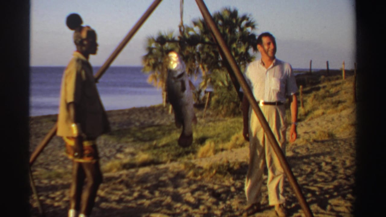 In this Sudan beach scene, the visitor faces the camera while the fisherman stands near a suspended catch and palm trees.