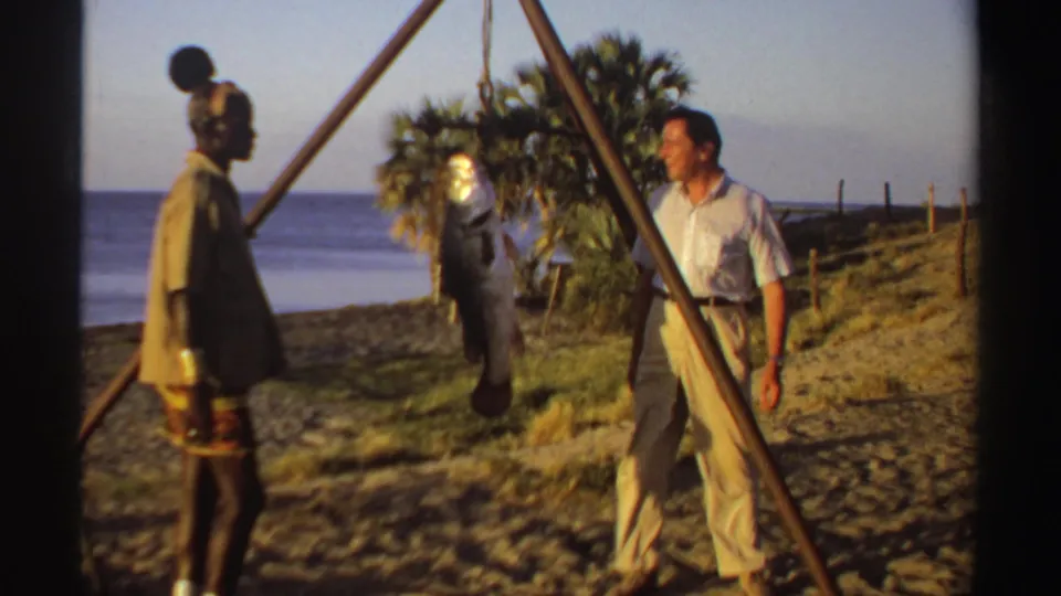 The visitor appears to look toward the fisherman and suspended fish in a 1969 Sudan coastal home-movie shot.