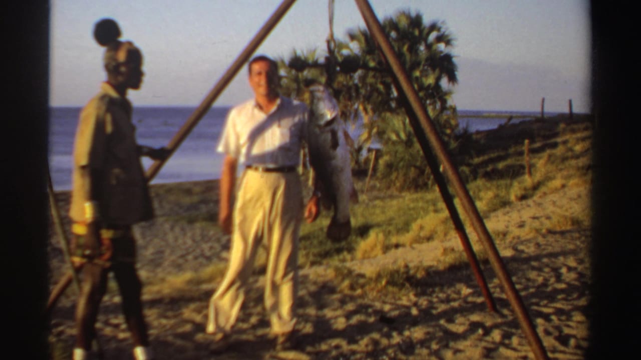 Two men stand on a sandy beach beside a large fish hanging from a wooden frame.