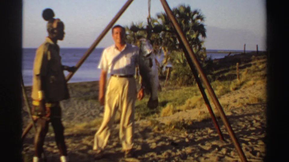 In 1969 Sudan footage, two adult men stand near the shore beside a large suspended fish and wooden tripod.