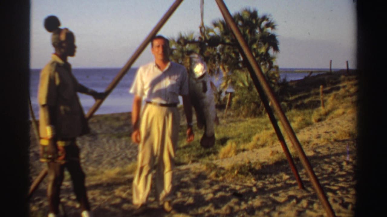 A white man faces the camera while another man stands near a hanging fish on the beach.