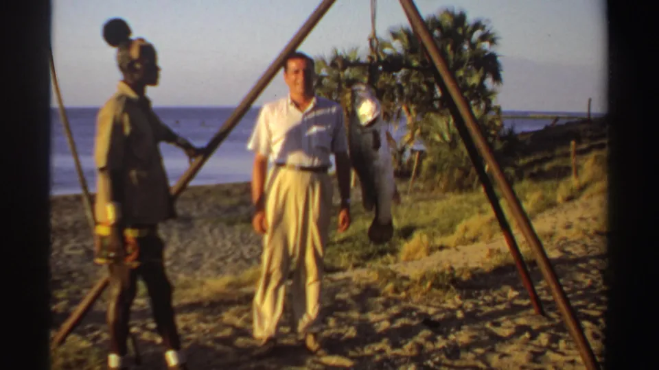 The 1969 Sudan home-movie frame shows two adults beside a hanging fish with palms and shoreline behind them.