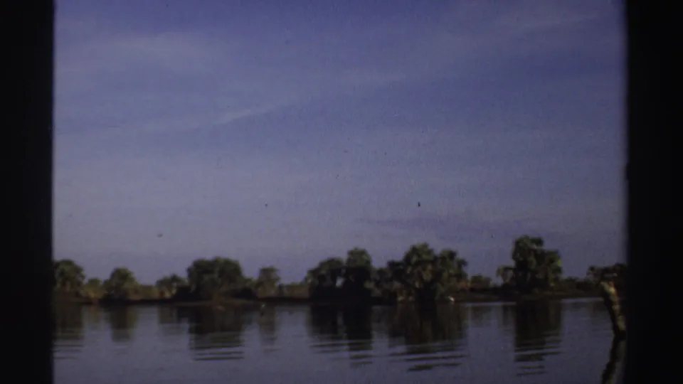 1969 Sudan 8mm frame looking across calm water toward a low tree-lined shore under an open sky.