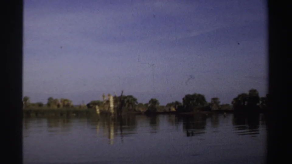 1969 Sudan 8mm frame showing a distant weathered stump in calm lake water, with trees along the far shore.