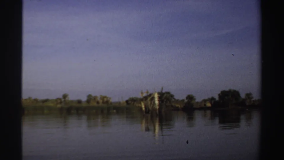 1969 Sudan 8mm frame showing calm lake water, a distant treeline, pale sky, and dark vertical film-frame edges.