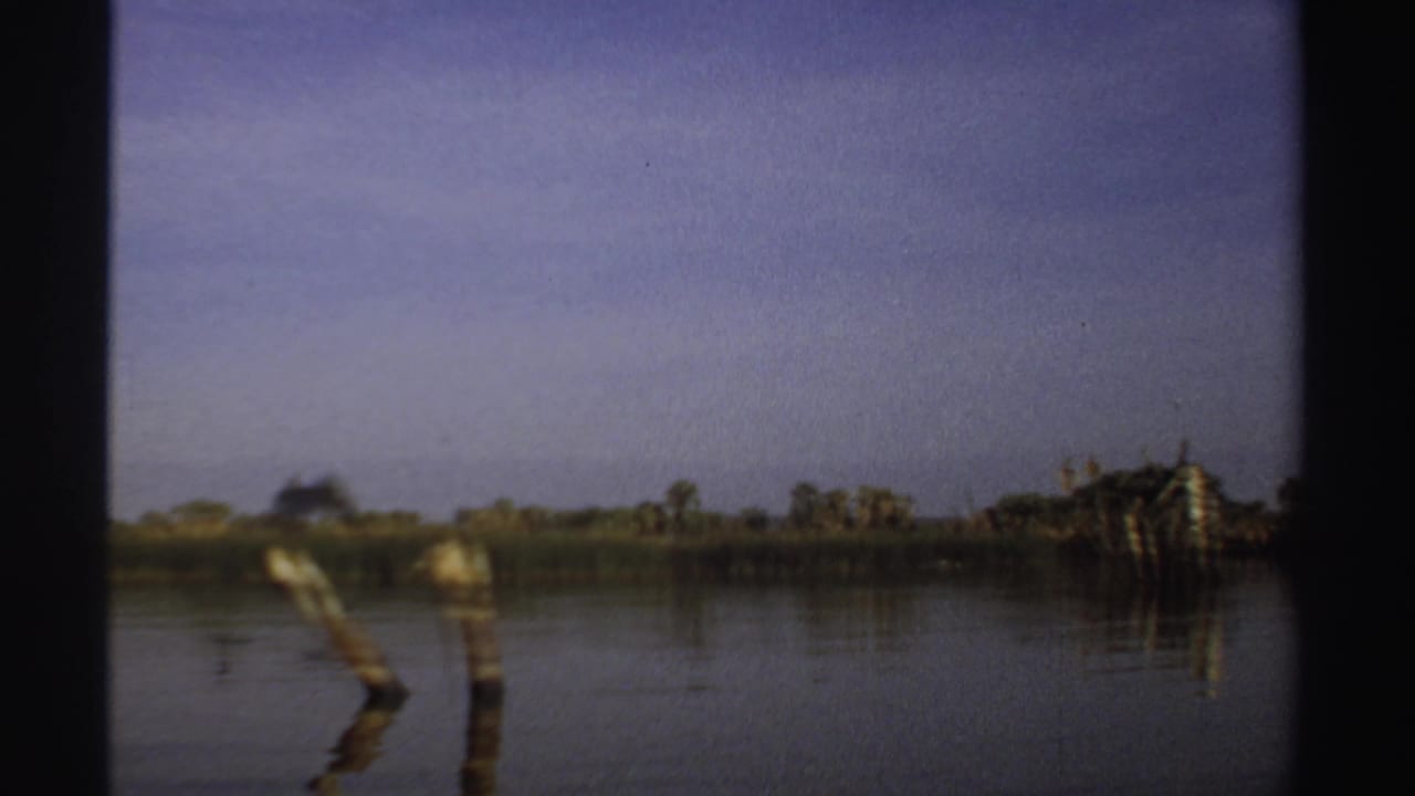 1969 Sudan 8mm frame with wooden posts rising from calm water, distant shoreline vegetation, and a pale blue-purple sky.