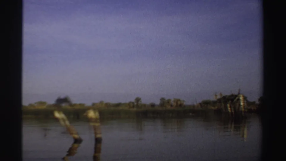 1969 Sudan 8mm frame with wooden posts rising from calm water, distant shoreline vegetation, and a pale blue-purple sky.