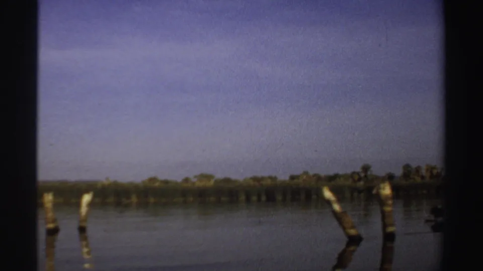 1969 Sudan 8mm frame showing a low lakeside horizon, wooden posts in the water, distant trees, and soft reflected light.