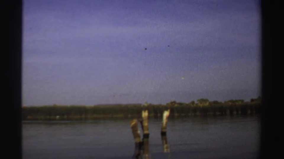 1969 Sudan 8mm frame with two wooden posts in still lake water, a thin distant treeline, and pale sky across the background.
